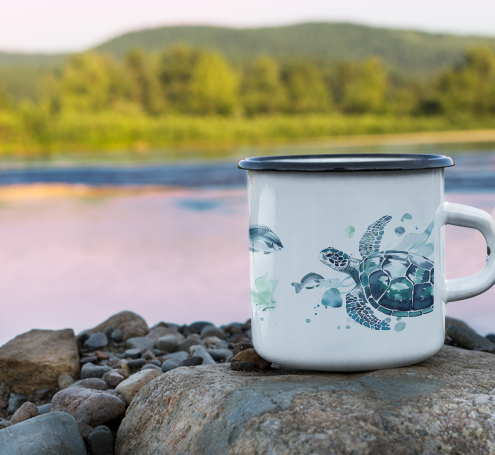 White enamel mug with a watercolour painting of a sea turtle and fish placed on the rocky shore of a lake